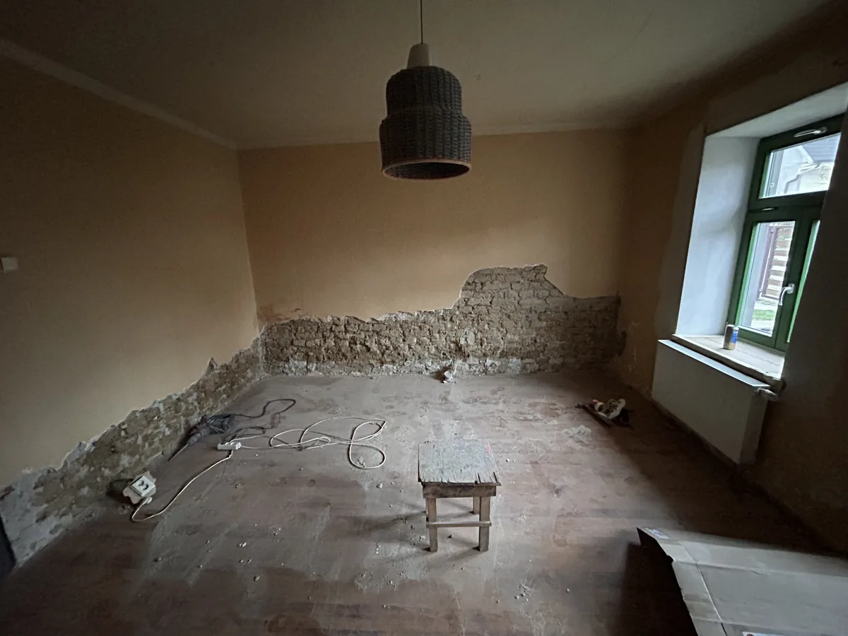Izba starého domu s odstránenou omietkou v dolnej časti stien, odkryté nerovné murivo z rôznych materiálov, drevená podlaha a okno so zeleným rámom